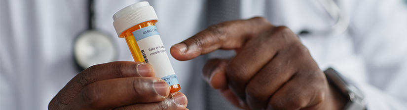 Middle aged Black man doctor holding prescription medication bottle, pointing at label with finger, wearing coat and stethoscope, demonstrating medication instructions